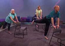 3 standing people holding backs of chairs and extending one leg